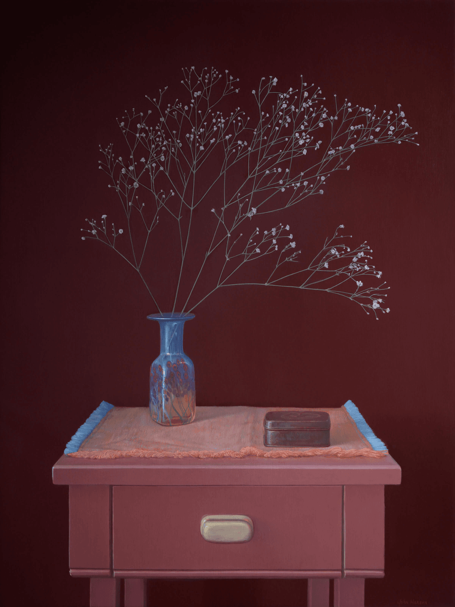 A realist still life oil painting titled 'Baby's Breath in a Glass Vase'. A pinkish-peach wooden side table with a single drawer stands against a solid, deep burgundy background. A fringed textile in peach and light blue rests on the table, supporting a translucent blue glass vase decorated with red floral motifs. The vase holds a sprawling spray of delicate baby's breath with tiny white blossoms. To the right of the vase sits a small, rectangular dark metal tin. The lighting is soft and even, emphasizing the clean lines of the furniture and the ethereal quality of the flowers. Oil painting by John Hansen.