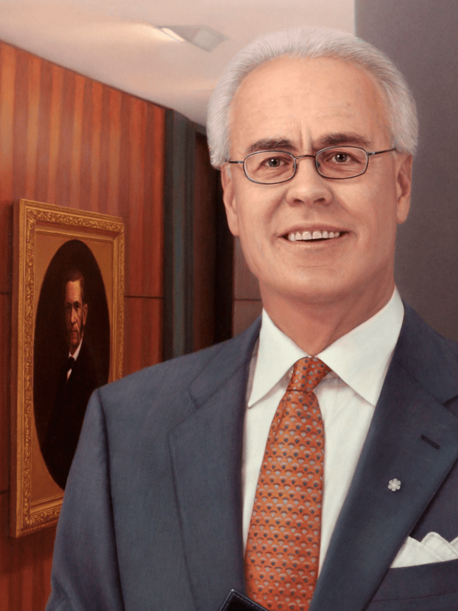 A detailed close-up from the oil painting 'Businessman'. This view focuses on the man's face and upper torso, showcasing the fine detail in his expression, the realistic reflections in his glasses, and the soft texture of his grey hair. It highlights the precision of the patterned tie, the small silver lapel pin, and the smooth, professional finish of the suit jacket. The background shows a portion of the wood paneling and the framed portrait in softer focus. Oil painting by John Hansen.