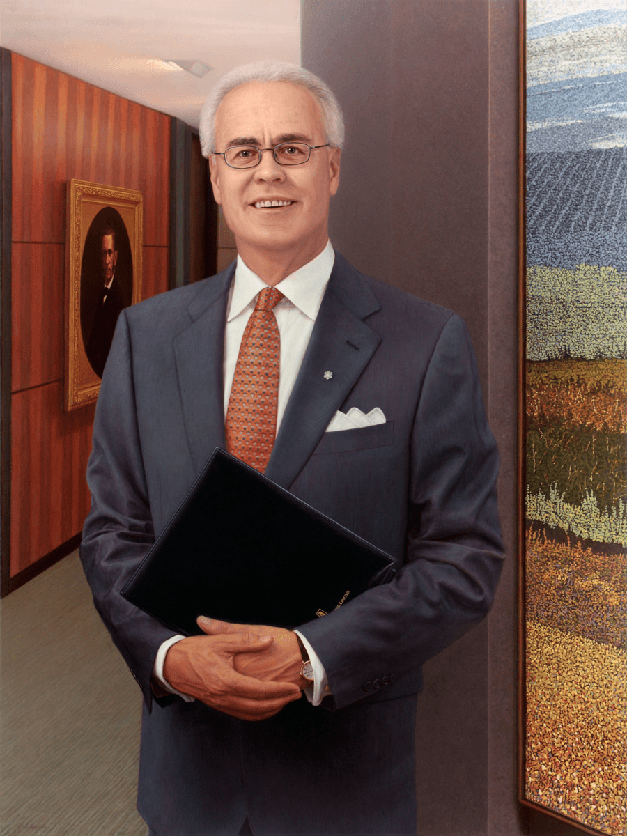 A formal realist oil portrait titled 'Businessman,' measuring 40 inches by 30 inches. A middle-aged man with short grey hair and spectacles stands in a modern office corridor, smiling directly at the viewer. He is dressed in a dark blue business suit with a white pocket square, a crisp white shirt, and an intricately patterned orange and red necktie. He holds a dark blue portfolio in front of him with both hands. The background features a wood-paneled wall with a gold-framed oval portrait to the left and a large, colorful landscape painting to the right. The lighting is bright and professional, highlighting the subject's friendly expression and the clean lines of the setting. Oil painting by John Hansen.