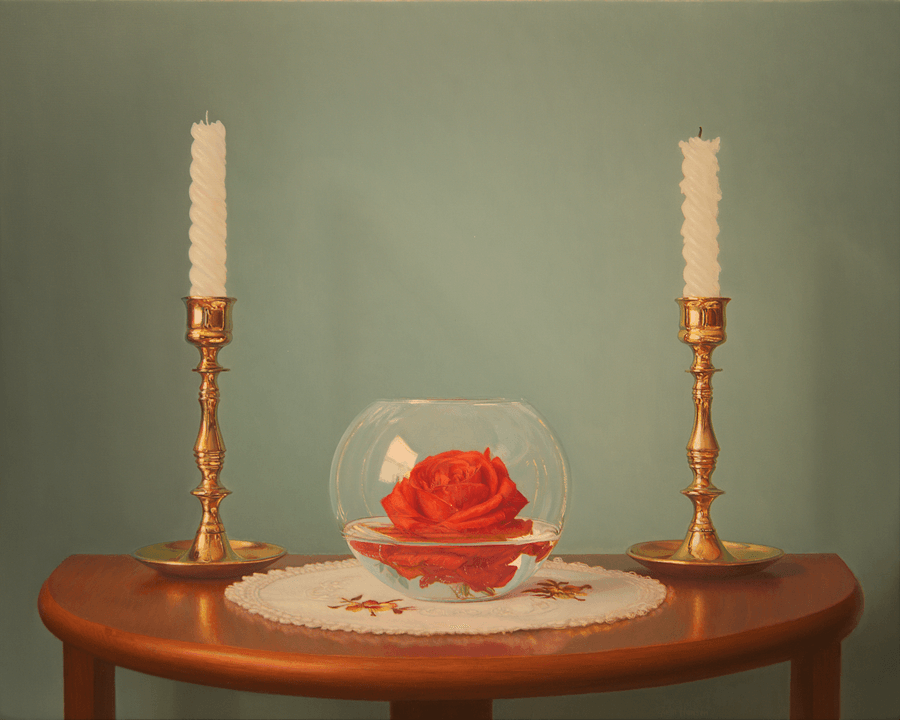 A realist still life oil painting titled 'Candles and Rose,' measuring 16 inches by 20 inches. Centered on a warm wood demi-lune table is a round glass bowl partially filled with water, in which a single, vibrant red rose floats. The bowl sits on a white lace doily embroidered with small yellow and orange flowers. Flanking the bowl are two tall, polished brass candlesticks, each holding a white, spiral-tapered candle. The background is a soft, muted teal-grey, and the lighting creates gentle highlights on the brass and a clear reflection of a window in the glass bowl. Oil painting by John Hansen.