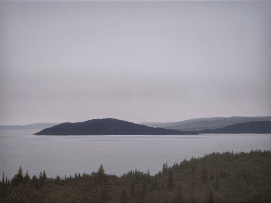 A serene landscape oil painting titled 'Lake Superior View'. A dense foreground of dark, finely detailed coniferous trees looks out over a vast, calm expanse of silvery-grey water. Several dark, forested headlands and islands sit in the middle ground, while layered hills recede into a misty, soft-purple distance under a flat, overcast sky. The muted palette and soft, diffused light create a quiet, atmospheric sense of northern vastness. Oil painting by John Hansen.