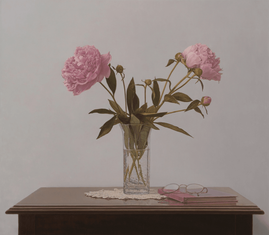 A realist still life oil painting titled 'Peonies,' measuring 20 1/2 inches by 23 1/2 inches. A crystal glass vase sits on a dark wood table, holding a lush arrangement of pink peonies with several unopened buds and deep green leaves. The vase rests on a delicate white lace doily. To the right of the vase, a pair of wire-rimmed glasses sits atop a closed, dusty-pink book. The background is a flat, neutral grey-white, and the lighting is soft and even, highlighting the intricate petals of the flowers and the reflections in the glass. Oil painting by John Hansen.