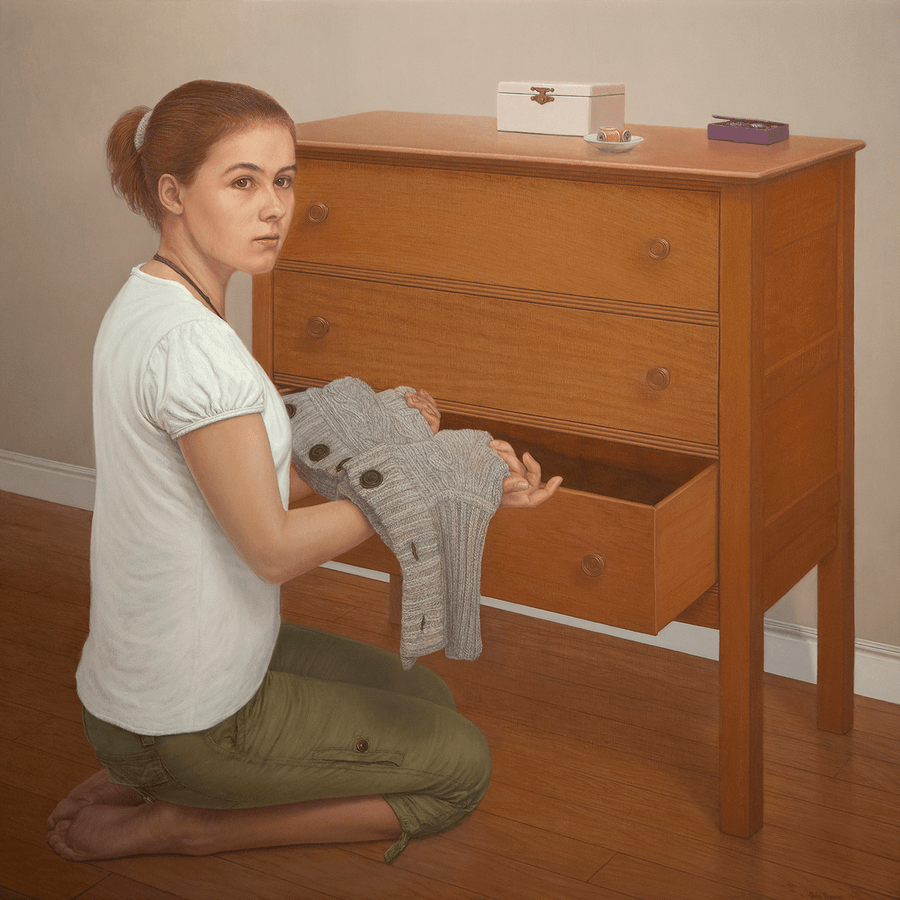A square realist oil painting titled 'Young Woman at Dresser,' measuring 24 inches by 24 inches. A young woman with reddish-auburn hair tied back is seen seated at a wood dresser. Her face is is turned in response to someone suggested within the room. She gently holds a heavily woven light grey sweater in her arms that has been taken out from an open drawer. The dresser top holds a few small items, including a jewellery box, a few spools of thread in a dish, and a small violet trinkets box that sits partially open. Soft, warm light from the left catches her shoulder and the warm surfaces of the wood, set against a light grey-green, muted interior background. Oil painting by John Hansen.