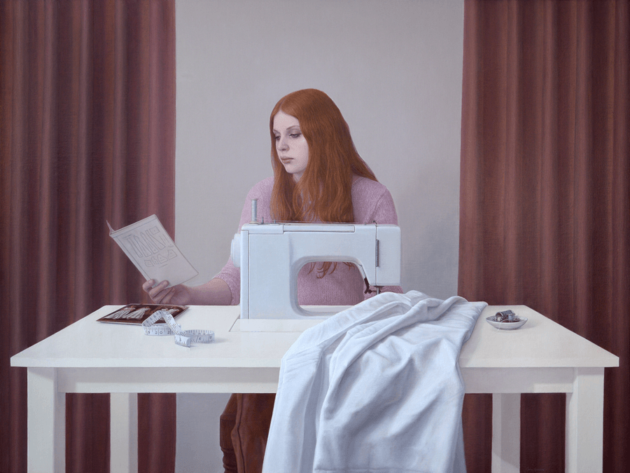 A realist figurative oil painting titled 'Young Woman at Sewing Table,' measuring 30 inches by 40 inches. A young woman with vibrant red hair is seated at a white wooden sewing table, her head bowed in concentration as she works on a piece of white fabric. Her right hand holds the sewing instructions for a dress. On the table sits a vintage white sewing machine, measuring tape, a small container, and spools of thread, including one bright red spool. The background is a quiet interior with a framed by earth red curtains. Soft, warm light from the left illuminates her profile and the sewing apparatus, creating an atmosphere of calm, steady industry. Oil painting by John Hansen.